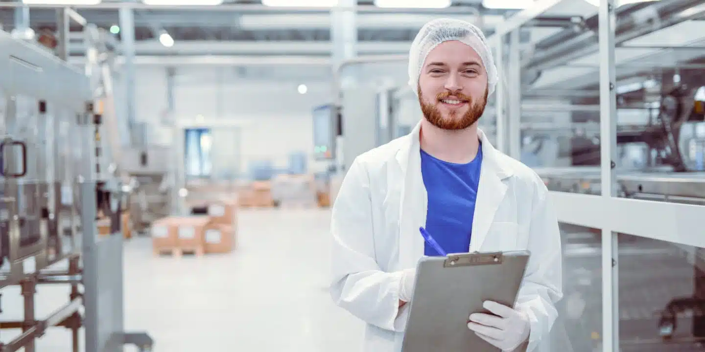 A man in a white lab coat and hairnet, focused on alternative proteins, holds a clipboard in an industrial setting with machinery.