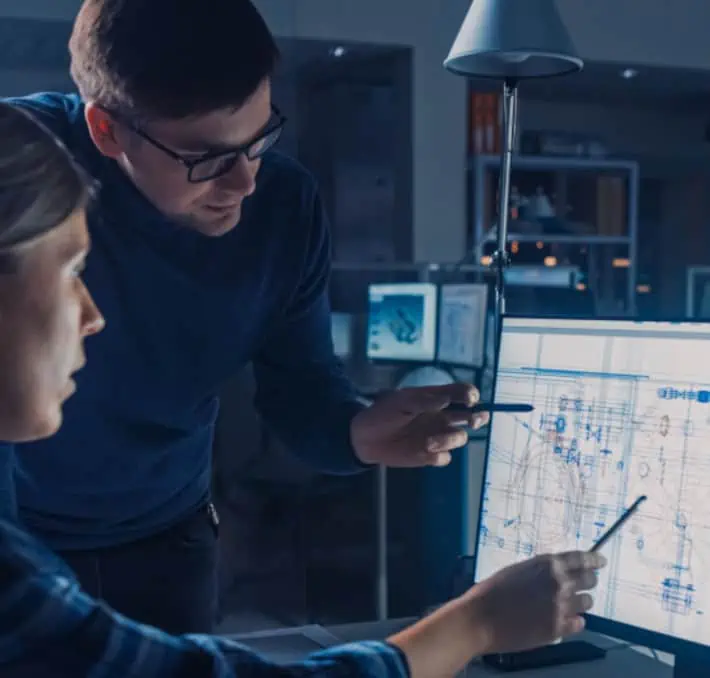 In a dimly lit office, two people intently discuss a blueprint on a computer monitor. One person sits, pointing at the screen displaying fluid equipment configurators, while the other stands beside them, gesturing thoughtfully with a pen.