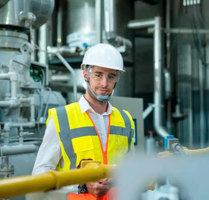 A worker in a reflective vest, hard hat, and safety glasses stands in an industrial setting with machinery and pipes, diligently monitoring fluid handling metrics for optimal performance.