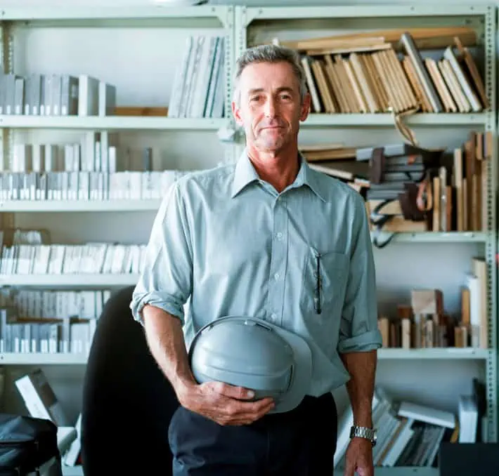 A CPQ administrator in a light shirt holds a hard hat in an office lined with shelves of books and materials.