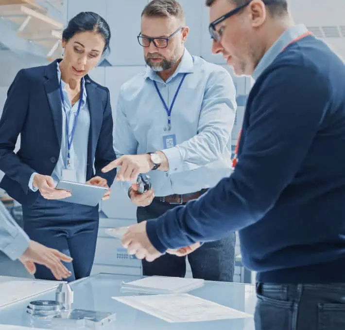 Three people in an office collaborate around a table with devices, papers, and blueprints.