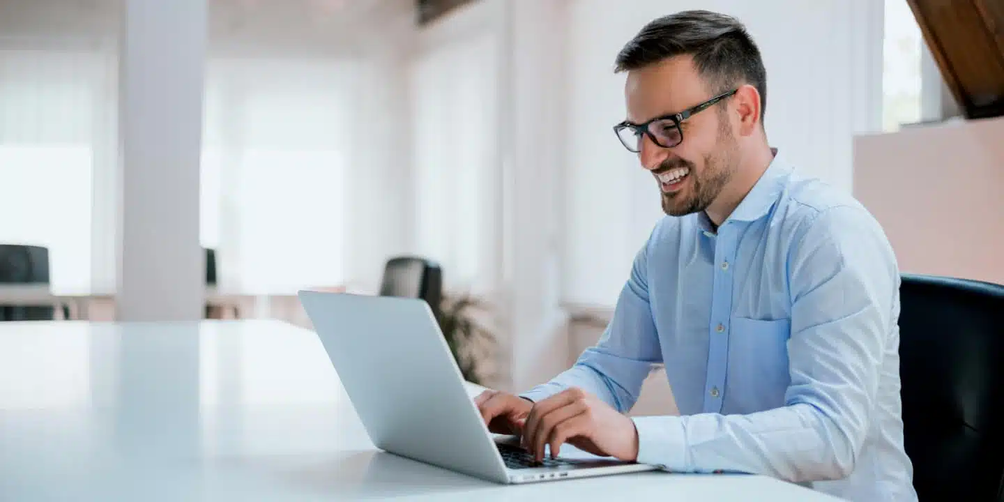 Man in blue shirt, smiling while typing on a laptop at a desk in a bright office, explores how to replace Excel with a product configurator.