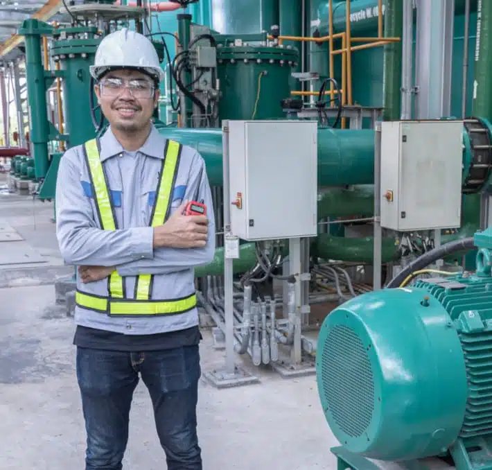 A person in a safety helmet and reflective vest stands with arms crossed in an industrial setting, surrounded by machinery, while closely monitoring fluid handling metrics in the background.