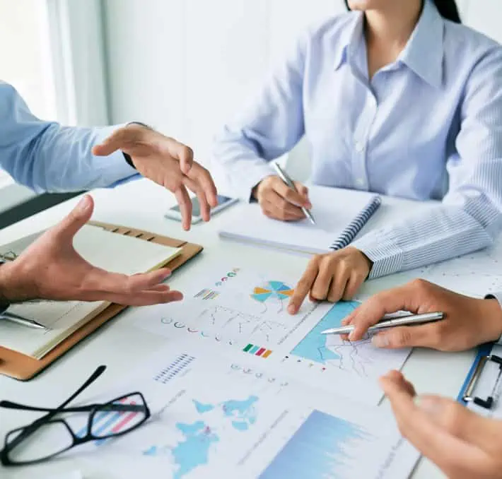 Three biomedical and clinical engineers are in a meeting room, discussing papers and charts. Hands gesture animatedly as they take notes and point at graphs on the table, analyzing data with precision.