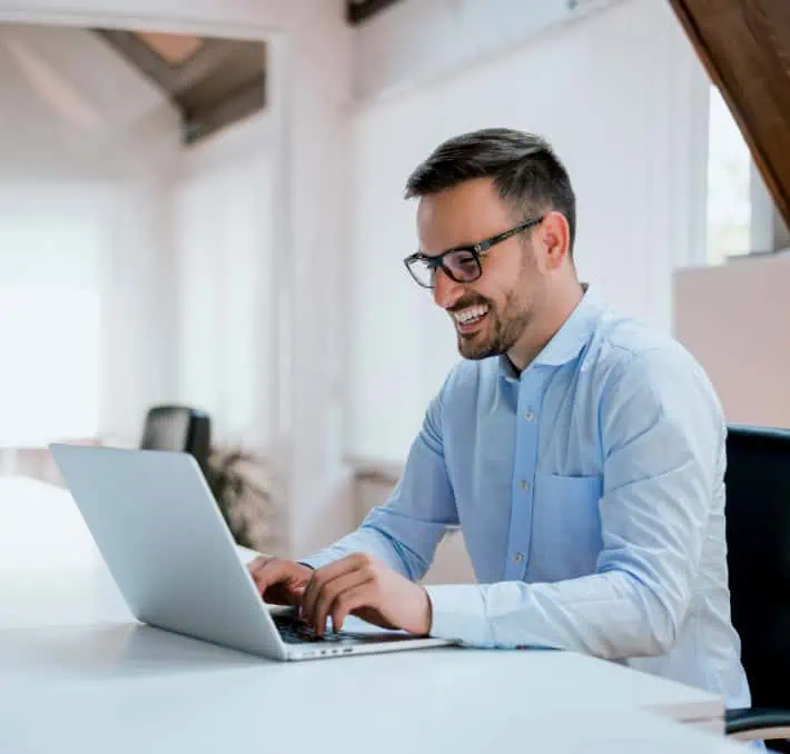Man in a light blue shirt, wearing glasses, smiles while typing on a laptop at a white desk. The office setting buzzes with B2B e-commerce activity, reflecting the digital ages dynamic business landscape.