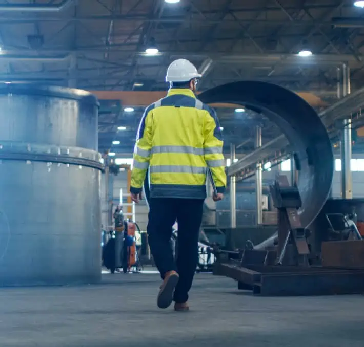 A person in a safety vest and hard hat walks through an industrial plant where CAD automation for manufacturing is seamlessly integrated into large equipment and metal structures.