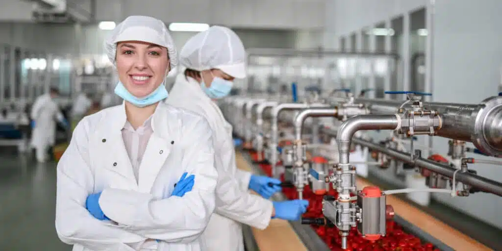 Workers in a food & beverage PLM plant. A woman in front is smiling, wearing a white coat and hairnet. Another worker focuses on machinery in the background, efficiently processing red produce.