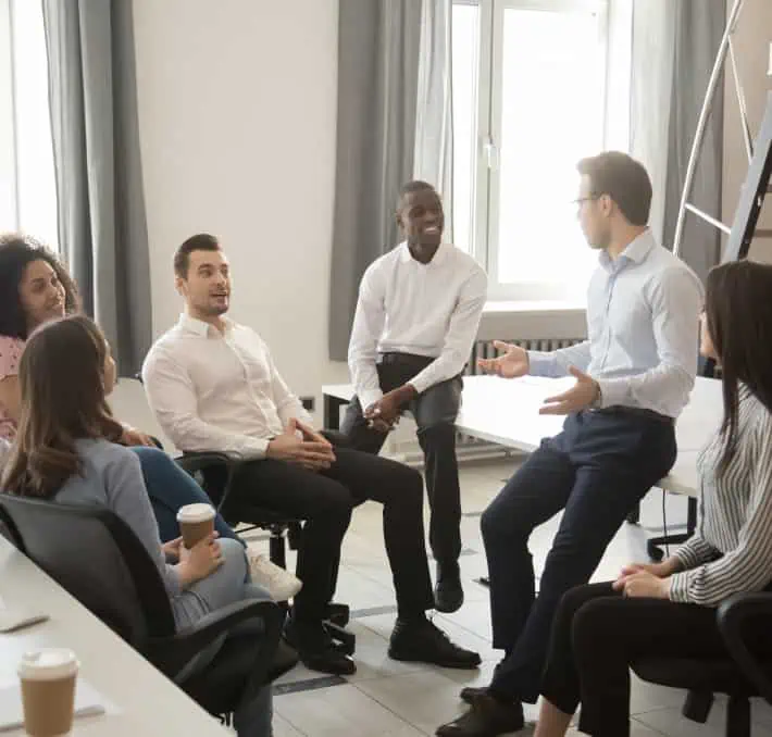 In an office setting, a group sits in a circle, engaged in conversation about emerging technologies. One person gestures animatedly while discussing the potential of digital twins for fluid simulations.