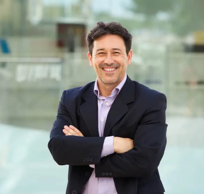 A man in a suit smiles confidently, arms crossed, embodying the perfect synergy of professionalism and innovation against a blurred office backdrop, hinting at the seamless integration of digital twin technology.