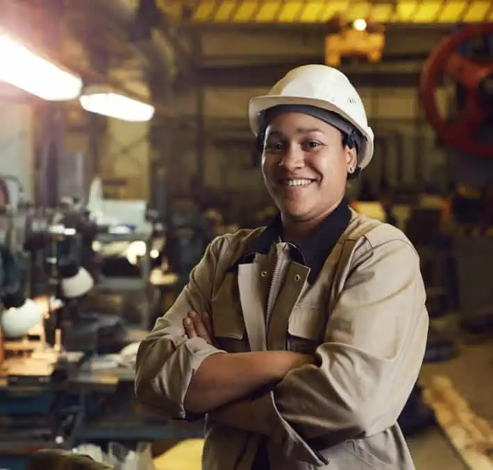 In an industrial workshop, a person in a hard hat and work clothes stands with arms crossed, smiling confidently. Their expertise in BOM Management ensures smooth operations and efficient production.