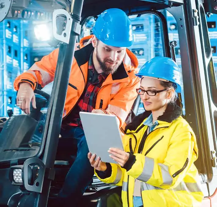 In a bustling warehouse, two workers in safety gear collaborate seamlessly: one sits confidently in a vehicle, while the other expertly navigates modern PLM solutions on a tablet, ensuring efficiency and precision.