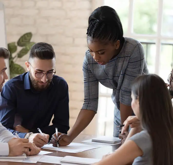 Een groep mensen in een vergadering, gericht op PLM-samenwerking, waarbij één persoon staat en naar documenten op een tafel wijst om de communicatie te verbeteren.