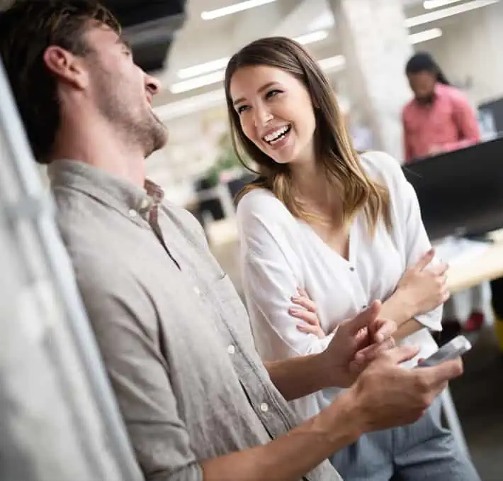 Two people are standing and laughing in an office setting, dispelling CPQ misconceptions. One holds a smartphone, perhaps sharing the latest industry insights. Other workers are visible in the background, engrossed in their tasks but occasionally glancing over with smiles.