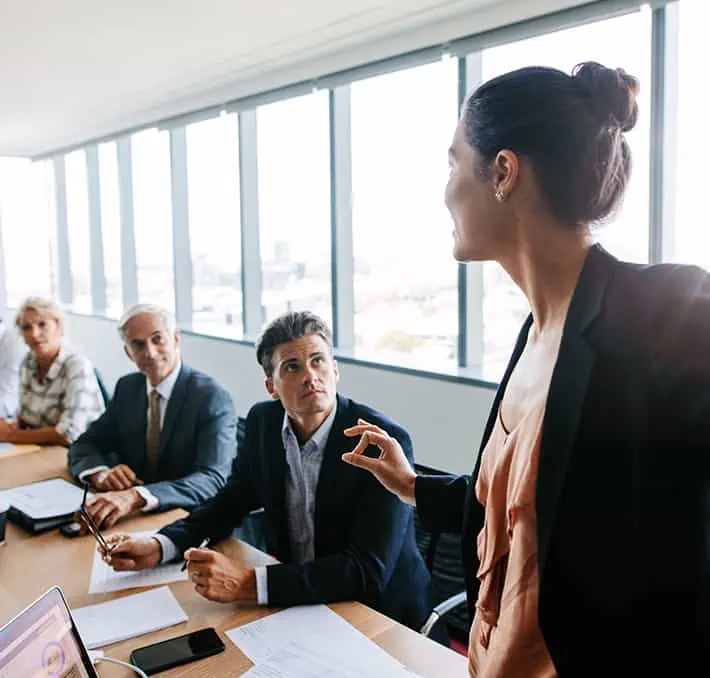 A woman in a suit gestures while discussing CPQ strategies with three seated colleagues in a conference room with large windows, highlighting the synergy with CRM systems.