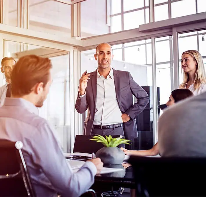 A man in a suit speaks confidently about CPQ and ERP systems to seated colleagues in a modern conference room.