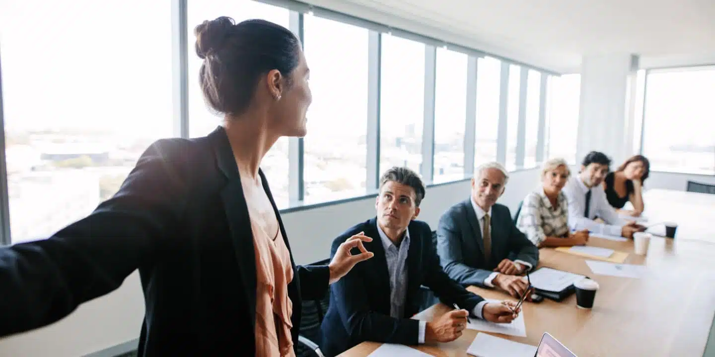 A woman stands and speaks to a group sitting at a conference table in a modern office, expertly addressing CPQ and CRM strategies.