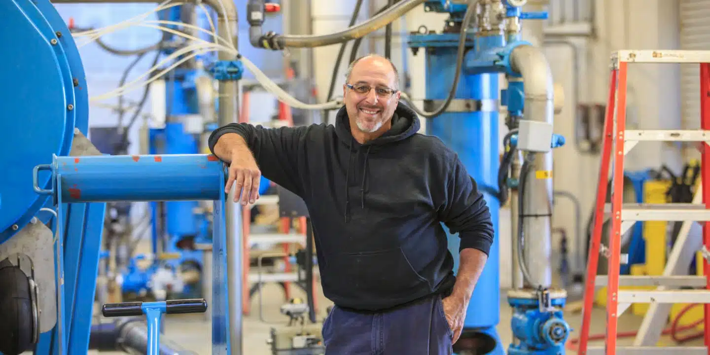 A man in a black hoodie smiles while leaning on blue industrial equipment in a workshop buzzing with the complexity of fluid process systems. A ladder stands in the background, surrounded by various intricate machines.