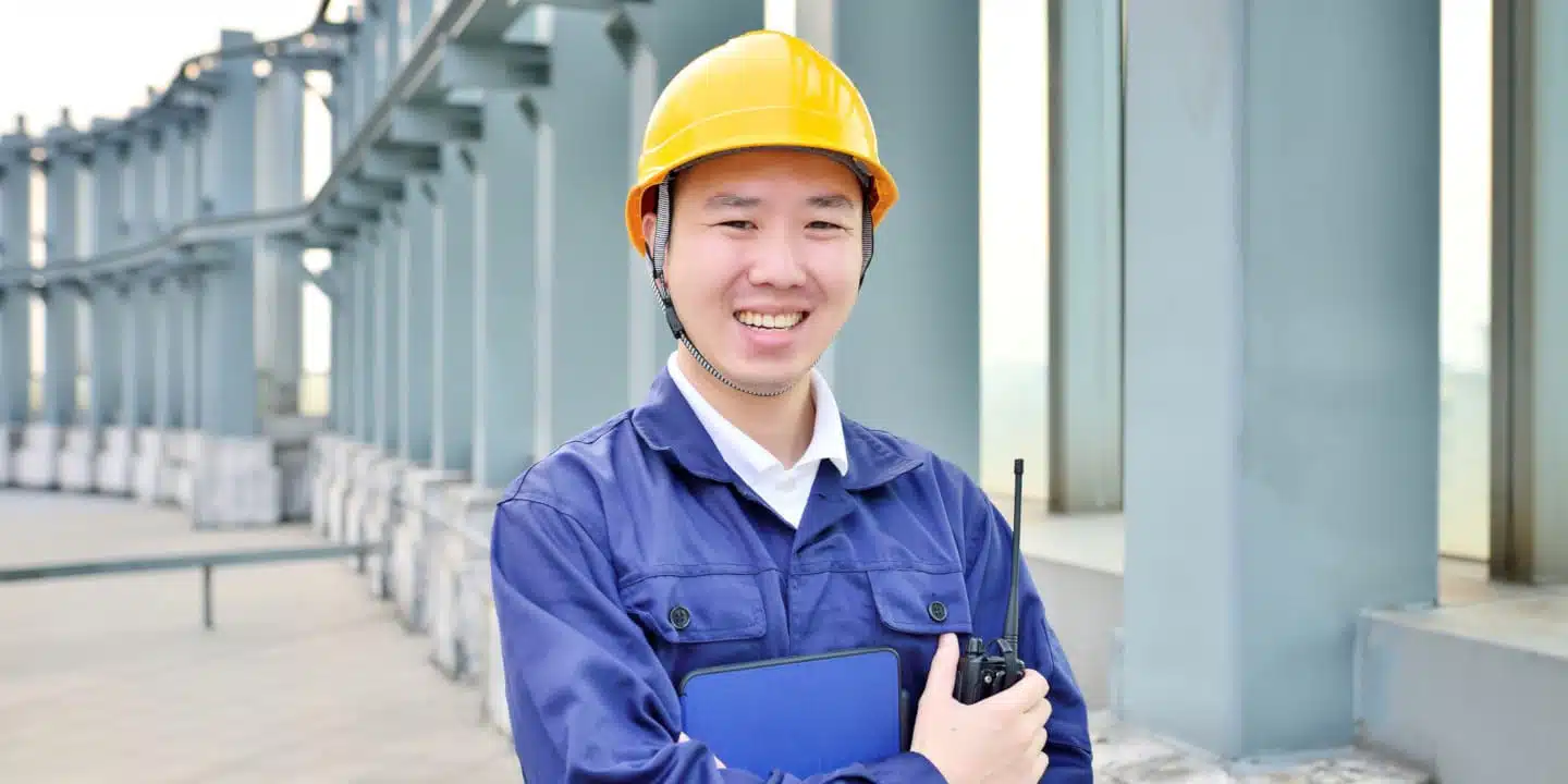 A person in a yellow hard hat and blue work uniform stands outside a building, smiling at the camera. With a clipboard ready for fluid flow calculations and a walkie-talkie in hand, theyre prepared for another successful day on the job.