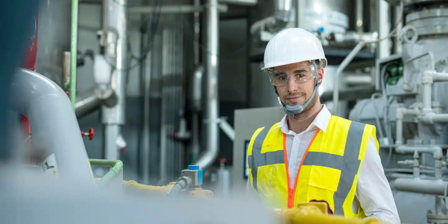 A man in a hard hat, safety glasses, and a yellow reflective vest stands in an industrial setting with machinery and pipes in the background, ensuring seamless fluid flow communication.