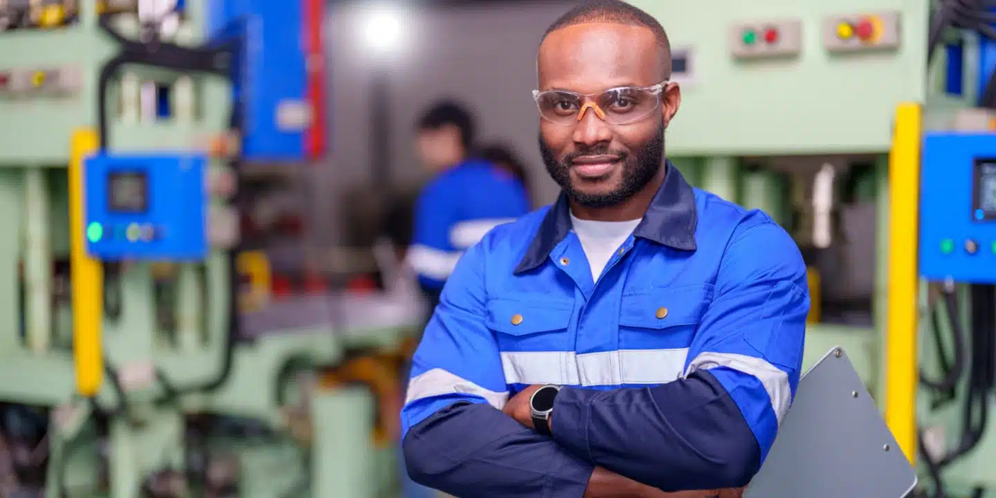 A person in safety glasses and a blue uniform stands confidently in an Industry 4.0 factory setting, holding a clipboard and crossing their arms. Advanced machinery hums alongside another worker diligently operating in the background.