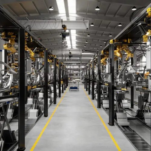 Interior of a modern factory with machinery and equipment aligned along a central aisle, under a high ceiling with skylights.