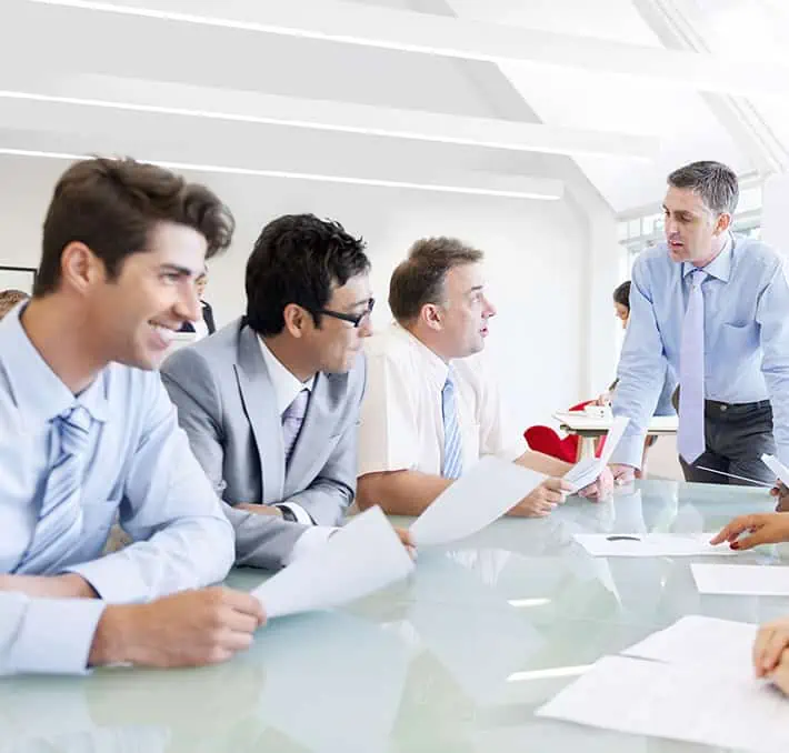 Five people in a meeting room, some seated and others standing around a table, are engaged in discussion. Dressed in business attire, they hold papers, efficiently guided by the Sofon Proposal Organizer to streamline their presentation materials.