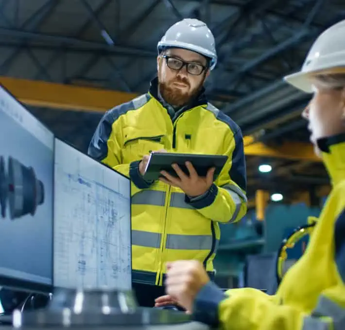 Engineer with a tablet observes a coworker working at a computer, focusing on 3D visualization in product configuration within the factory setting. Both wear safety gear as they collaborate on intricate technical drawings.
