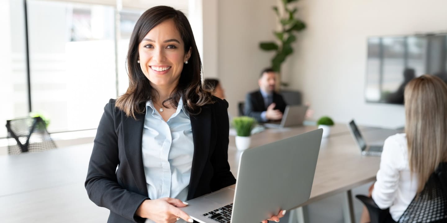 A woman in a professional setting holds a laptop and smiles confidently, perhaps after finalizing BOMs in electrical equipment. In the background, three colleagues sit at a table in a meeting room.