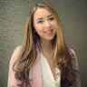 A person with long brown hair wearing a pink blazer and white top smiles in front of a textured wall.