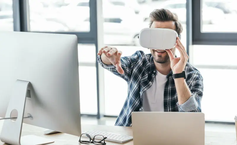 A man wearing an AQ Visual virtual reality headset interacts seamlessly with a virtual environment, fully immersed while seated at his desk equipped with both a computer and laptop.