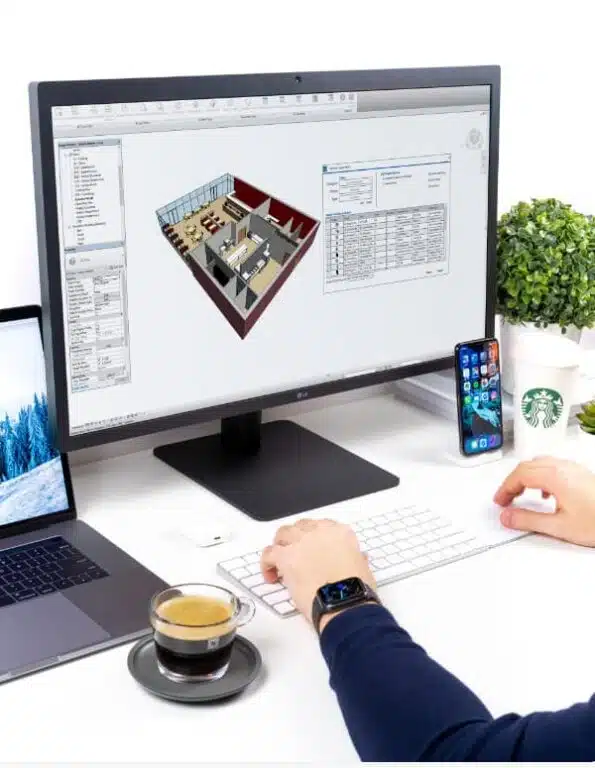 A person at a desk engrossed in architectural software on their desktop, with the laptop showcasing foodservice equipment design solutions nearby. A phone, cup of coffee, and plant add to the workspace ambiance.