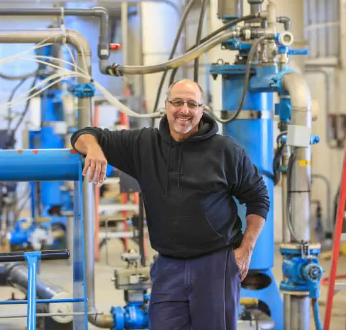 In an industrial setting filled with blue machinery and pipes, a person in a black hoodie stands smiling, embodying the spirit of innovation perhaps sparked by breakthroughs in fluid modeling software.