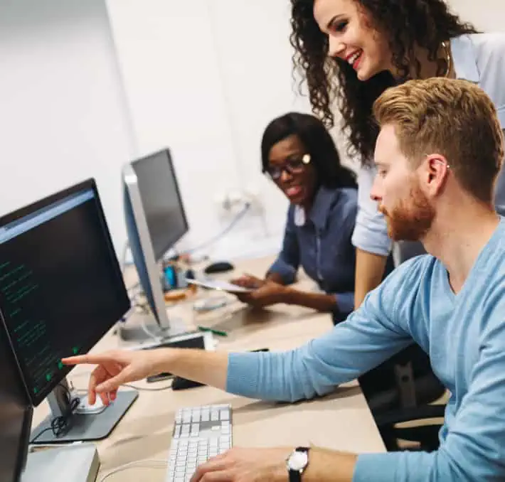 Three people at a desk work on computers, with one person pointing at a screen displaying code from AutoQuotes Insight Pro.