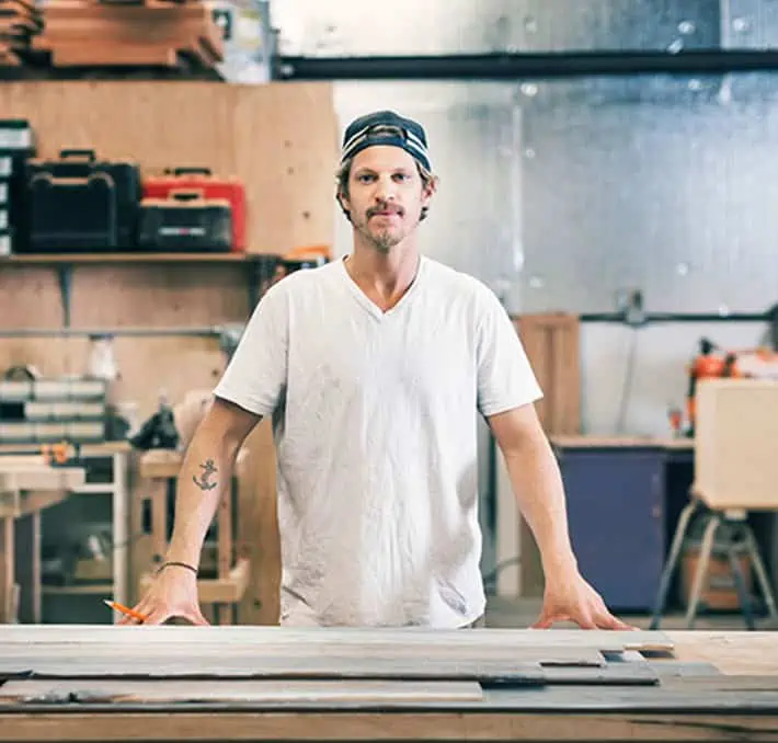 Person in a white shirt and backward cap stands at a workbench in a workshop with tools and wood in the background.