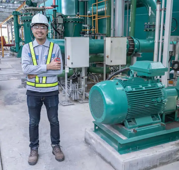 A worker in safety gear stands beside large industrial machinery in a factory setting, efficiently overseeing operations through the Intelliquip Project and Quote Management Portal.