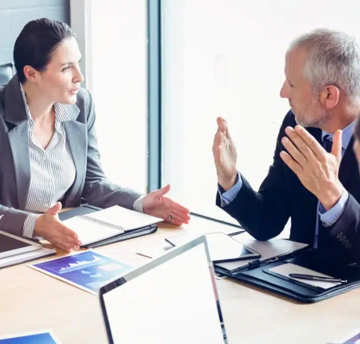 Two people in business attire engage in a discussion at a table with notebooks and reports, near a large window. They explore how to configure the SAP CPQ system effectively, while a laptop sits in the foreground, ready to support their cloud-based solutions.