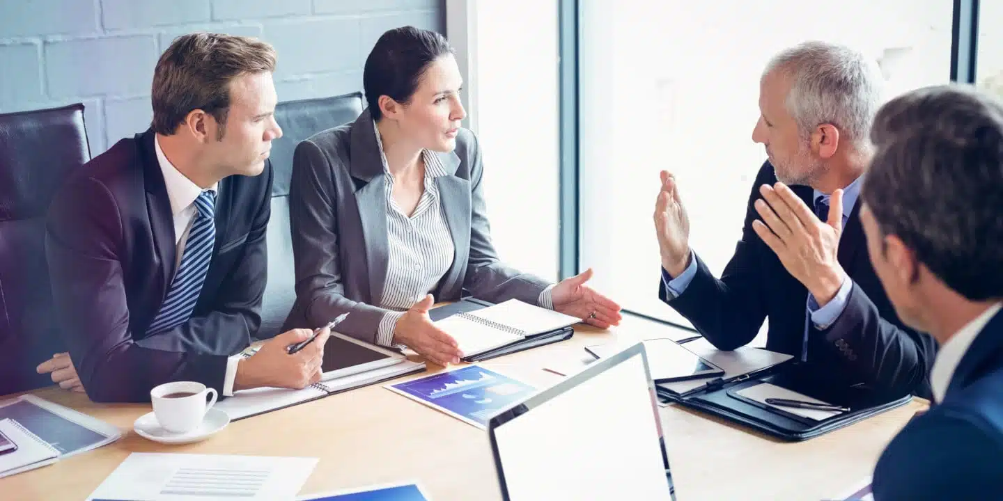 Four business professionals in a meeting room engage in a discussion about configuring One Cloud SAP CPQ. They sit around a table equipped with laptops, documents, and steaming coffee cups, tackling strategic decisions to enhance their cloud solutions.