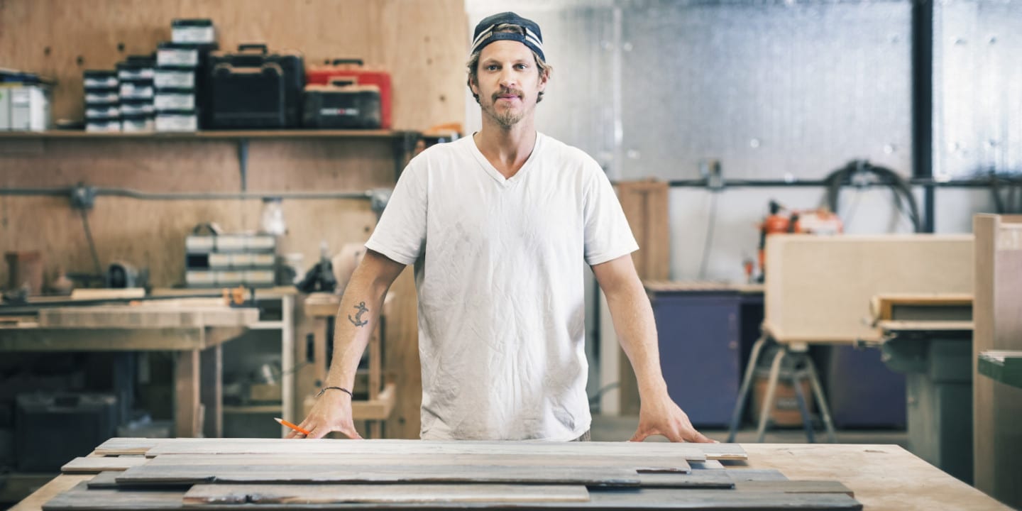 A man in a white t-shirt stands at a workbench in a woodworking shop, surrounded by tools and wooden boards, envisioning innovative furniture website features to showcase his handcrafted pieces.