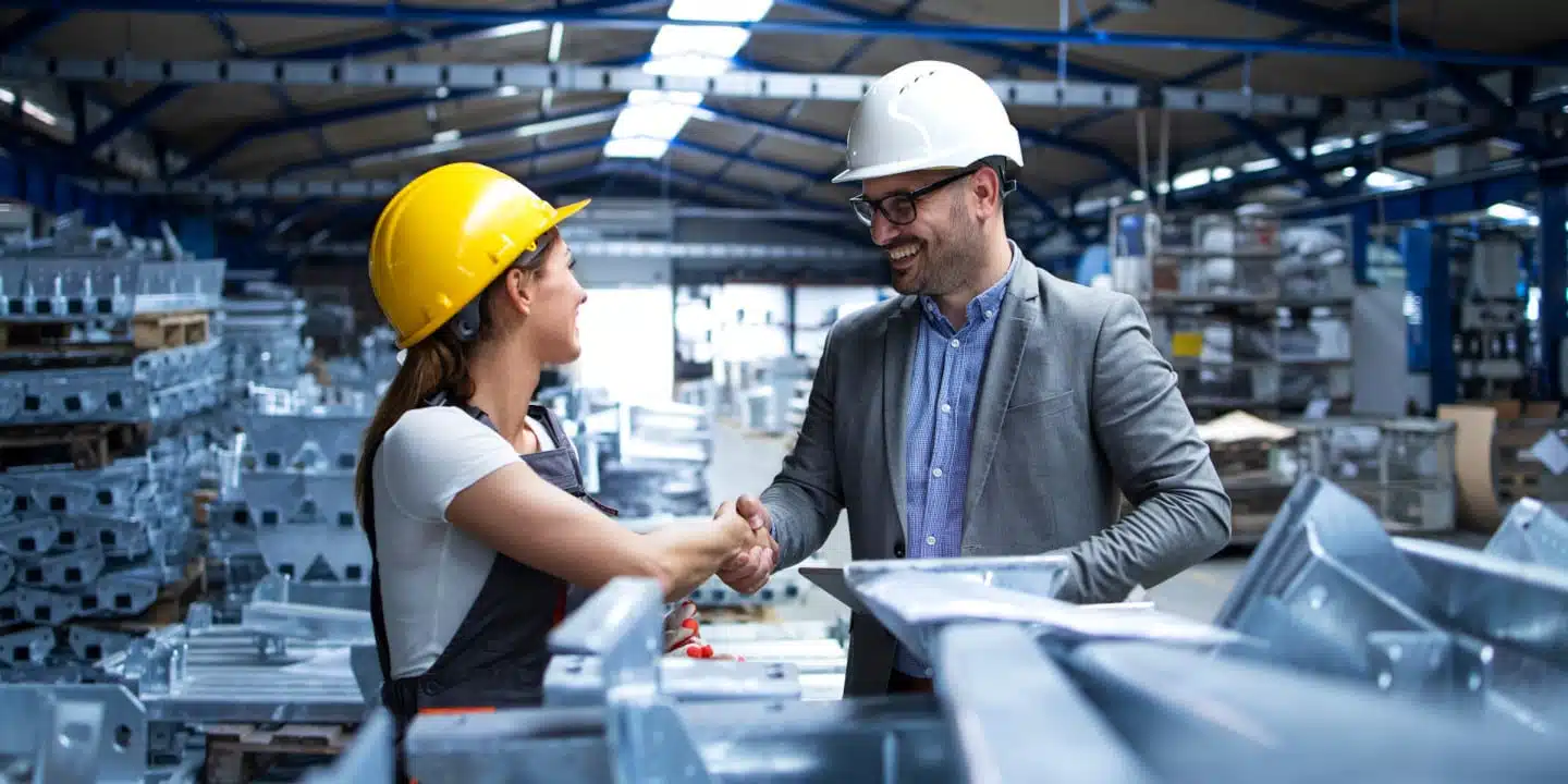 Two people wearing hard hats shake hands in a factory dedicated to instrumentation manufacturing, surrounded by industrial materials and metal beams.