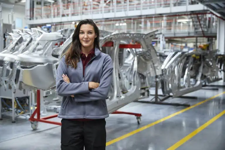 A person stands confidently with arms crossed in a BLEICHERT car manufacturing facility, surrounded by metal car frames and industrial equipment.