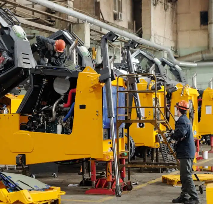 Under bright lighting, workers in a factory meticulously assemble large yellow industrial machinery with advanced lifting mechanisms, demonstrating the sophisticated CPQ for construction equipment manufacturing.
