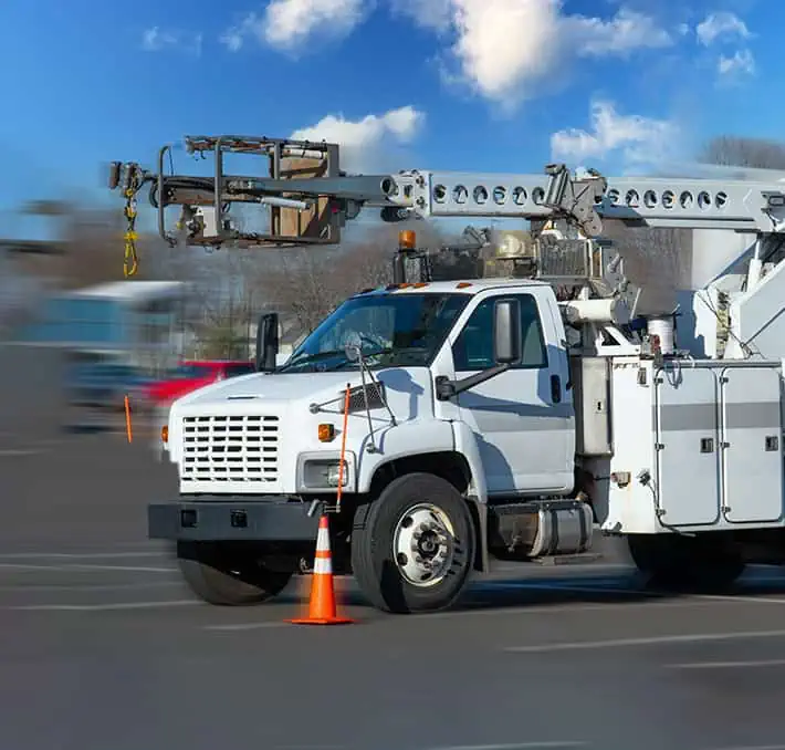 A white utility truck, crafted with excellence in specialty vehicle manufacturing, features an extended boom lift and is parked beside an orange traffic cone on a paved surface under a blue sky dotted with clouds.