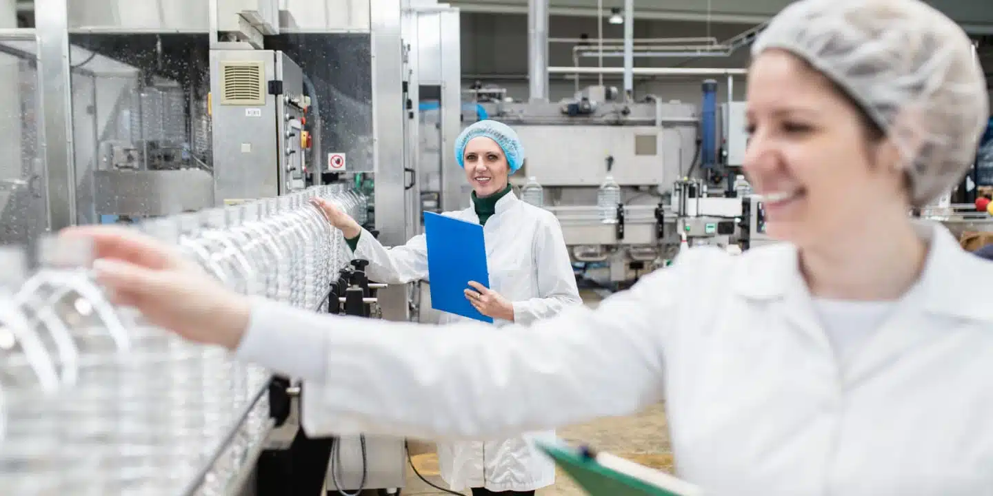 Workers in a bottling plant meticulously inspect plastic bottles on a production line, ensuring food & beverage compliance. Dressed in hair nets and white lab coats, they hold clipboards, diligently documenting their findings.