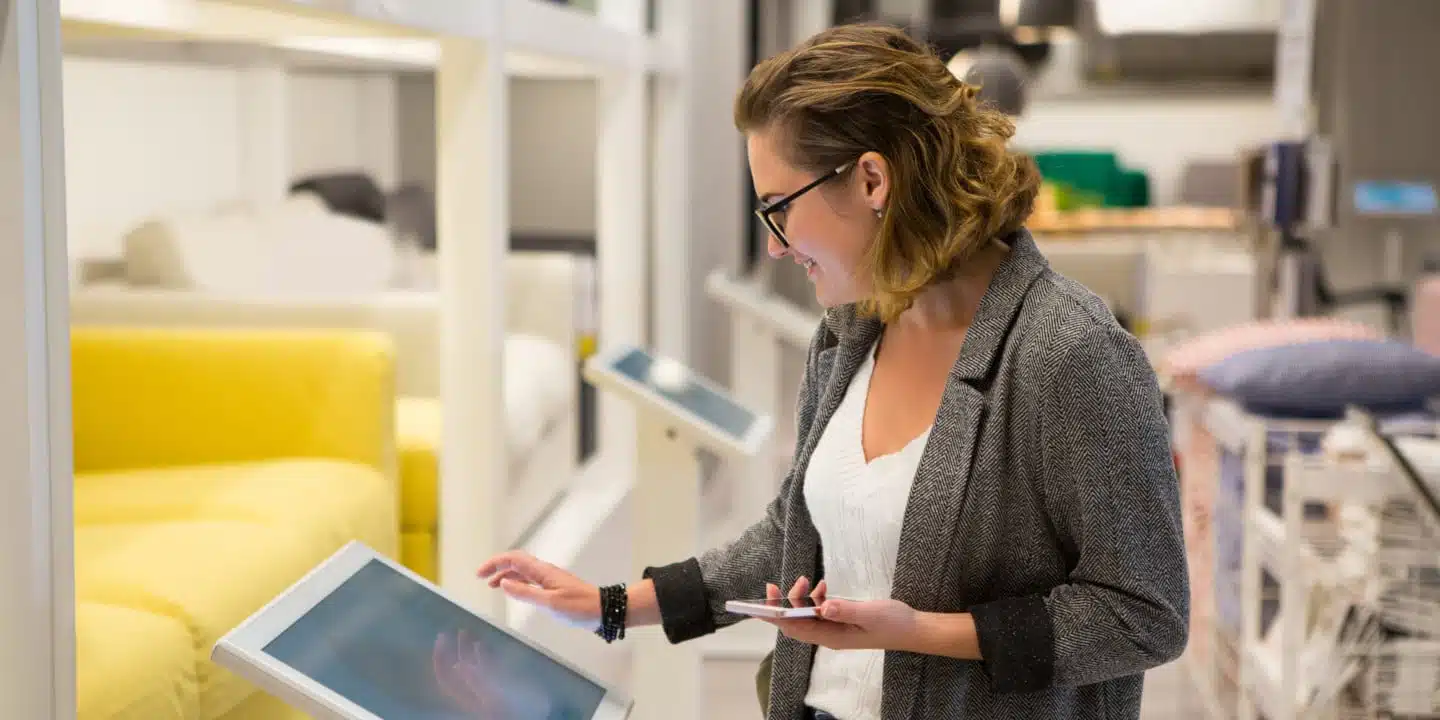 A woman in glasses and a grey blazer engages with a touchscreen in a modern showroom, reflecting on the latest marketing trends for furniture retailers, with stylish sofas set elegantly in the background.