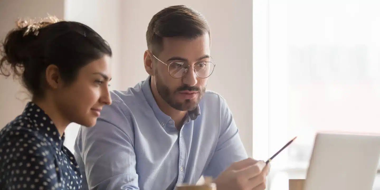 Two people seated at a desk focus on a laptop screen. A bearded person wearing glasses gestures with a pen, discussing CPQ software adoption, while the other listens attentively.