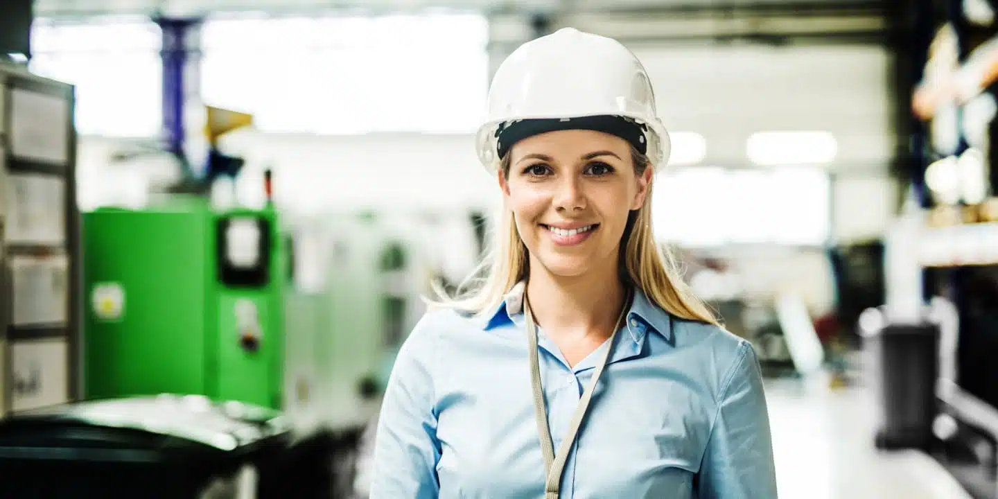 In an industrial setting, a person wearing a white hard hat and blue shirt stands confidently, showcasing the smile of someone adept in CPQ for Elevator and Moving Stairway Manufacturing.