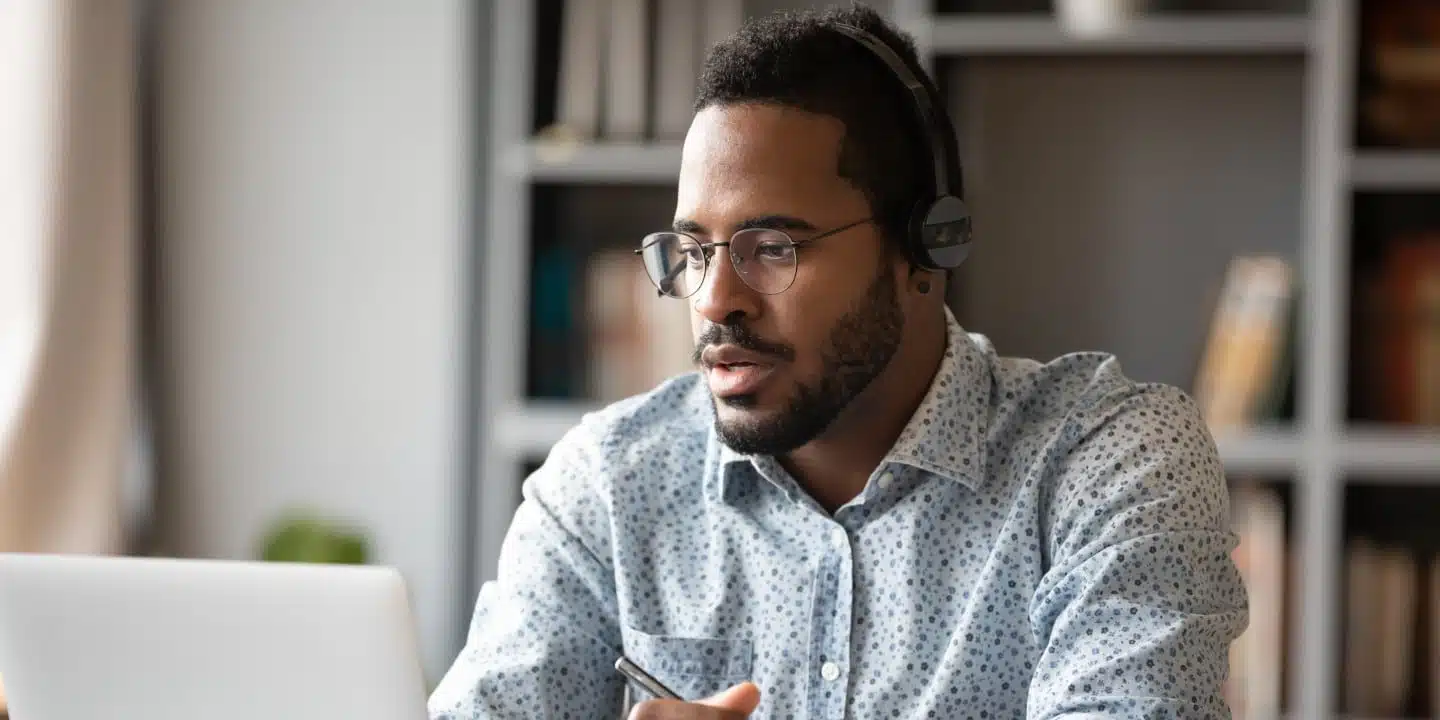 A person wearing glasses and headphones is seated at a table, using a laptop in a room with bookshelves filled with PLM and ERP solutions in the background.