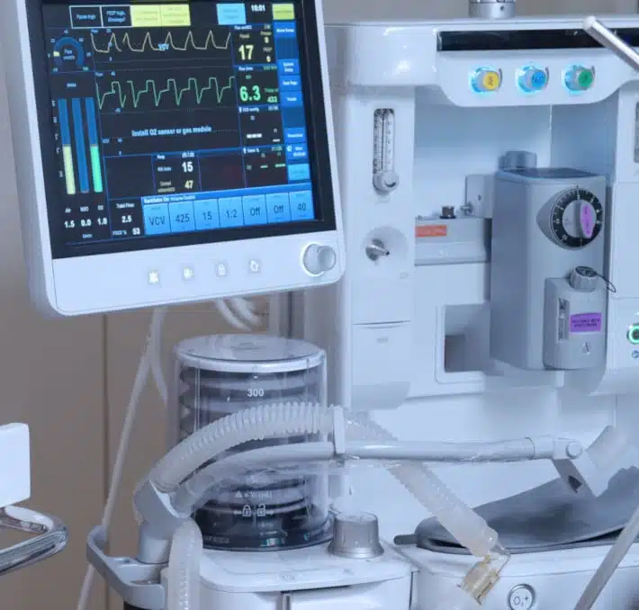 Close-up of biomedical devices, including a monitor displaying vital signs and a ventilator with attached tubes and controls, in a clinical setting.