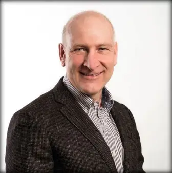 A man in a dark blazer and striped shirt smiles against a plain white background, exuding confidence—much like the detailed Prozesse zur Spezifikation von Lebensmitteln und Getränken that ensure quality.