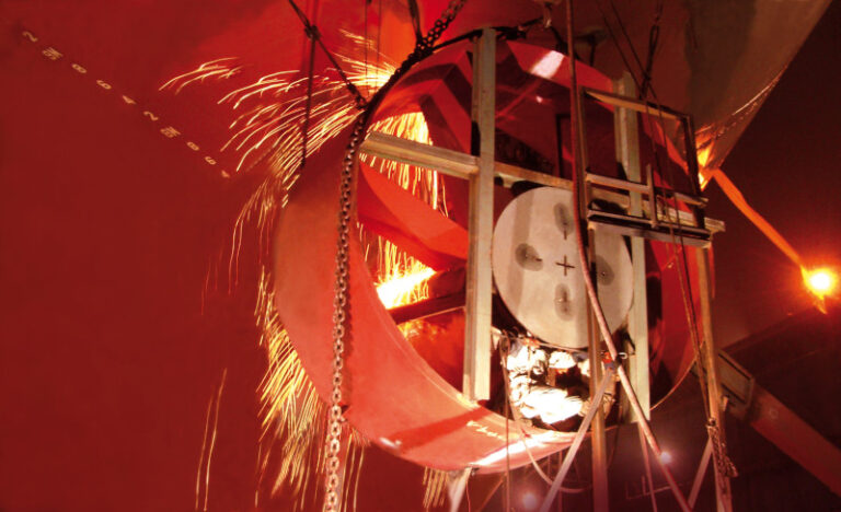 A circular structure with chains and sparks flying, reminiscent of Becker Marine Systemes robust designs, is illuminated in a dark setting, suggesting industrial or construction activity.
